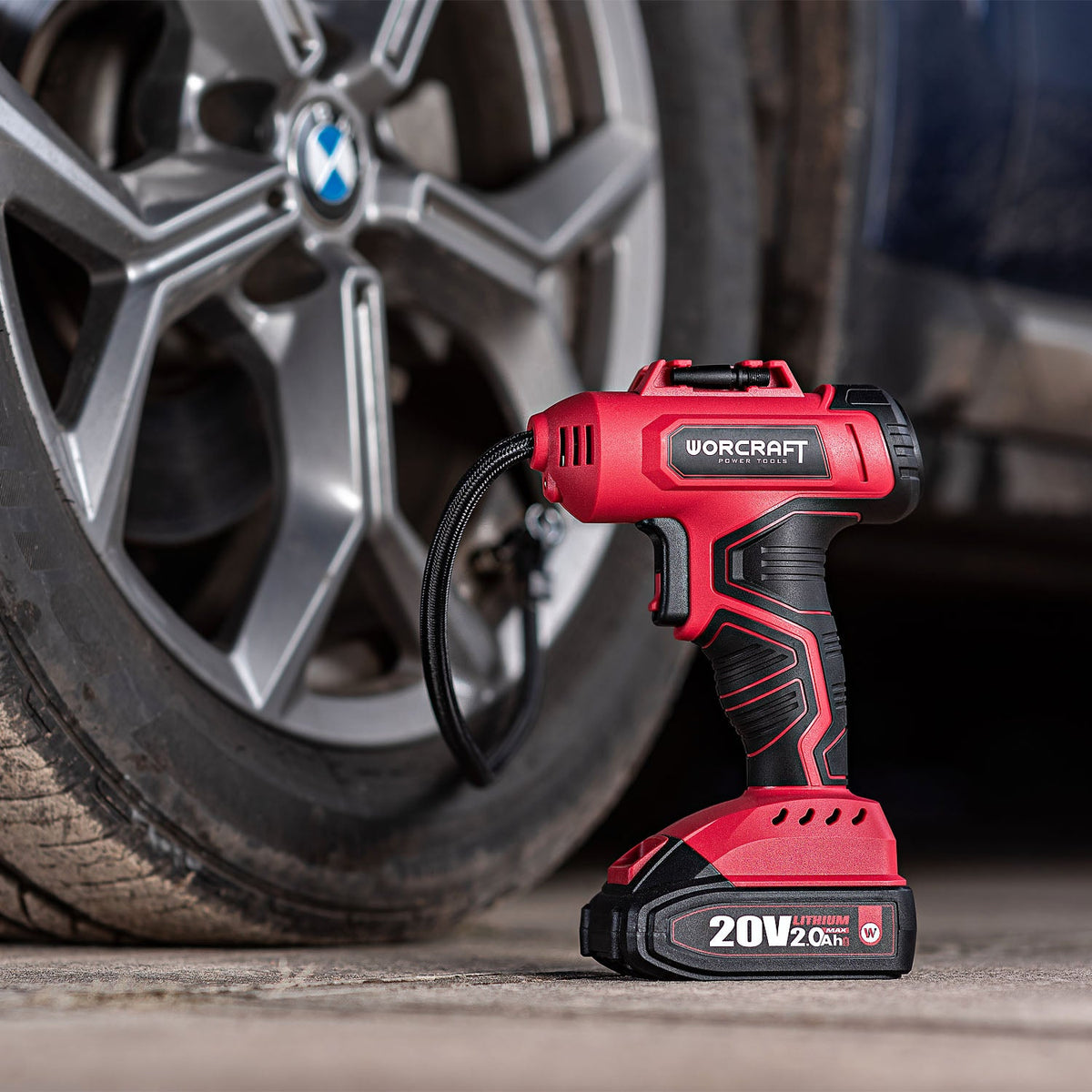 A red and black WORCRAFT cordless tire inflator standing on a concrete surface in front of a blurred car wheel.