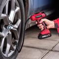 A hand holding a red and black worcraft cordless air pump tool connected to a car tire valve, shown in a real-world garage setting.