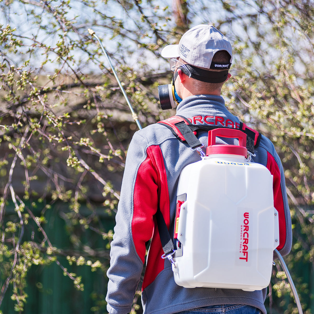 A man wearing a gray and red work jacket, a white cap, and a respirator mask is shown from behind, carrying a white WorCraft backpack sprayer outdoors among leafy trees.