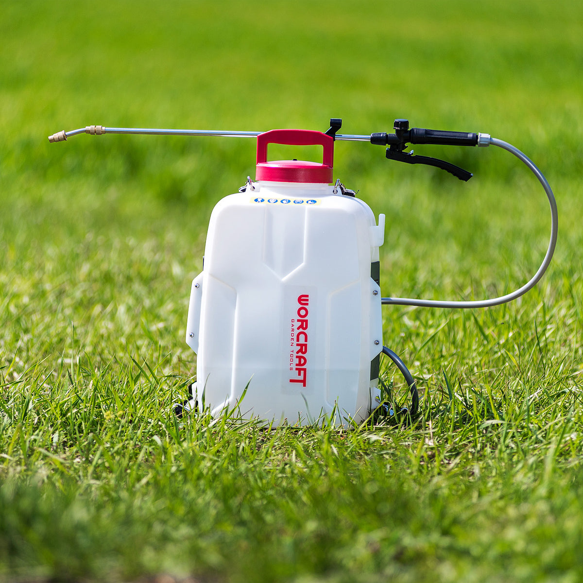 A white Worcraft backpack sprayer with a red cap and a long metal spray wand, set on a green grass lawn.