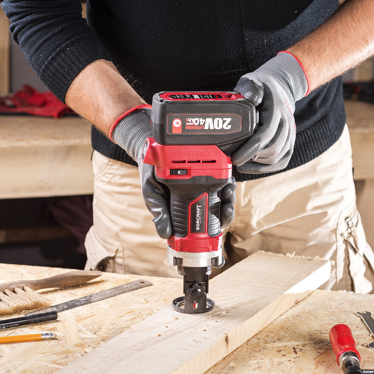 A person wearing gray gloves uses a red and black Worcraft 20V MAX cordless router to cut a piece of wood on a workbench.
