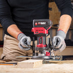 A person wearing gray work gloves uses a Worcraft red and black cordless router with a plunge bit on a piece of wood in a workshop setting. The router has a black handle with red accents and a visible battery pack labeled 20V MAX.
