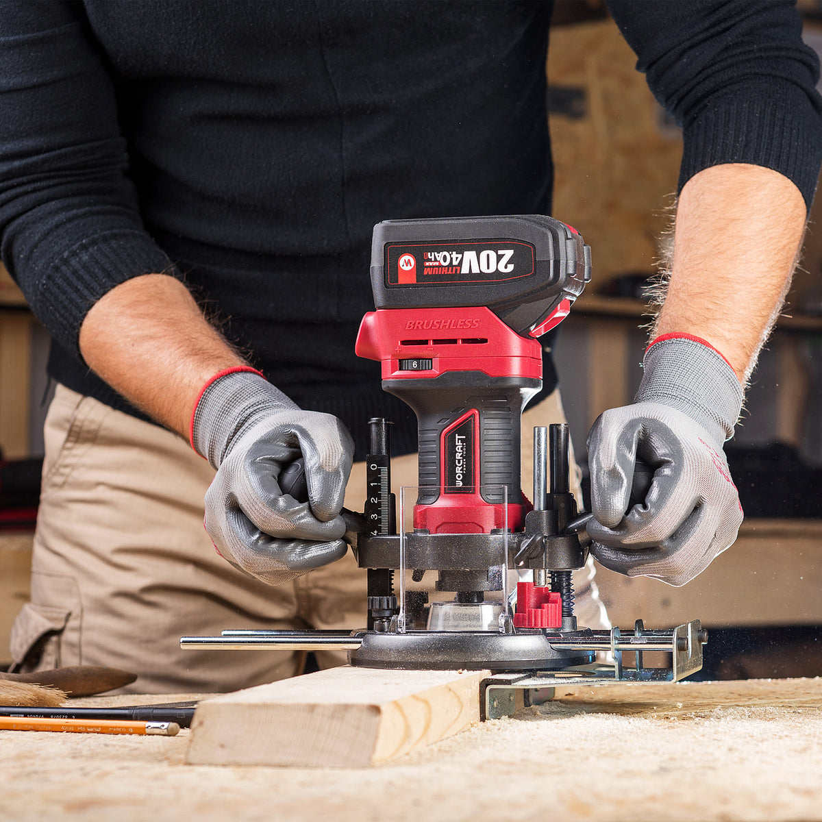 A person wearing gray work gloves uses a Worcraft red and black cordless router with a plunge bit on a piece of wood in a workshop setting. The router has a black handle with red accents and a visible battery pack labeled 20V MAX.