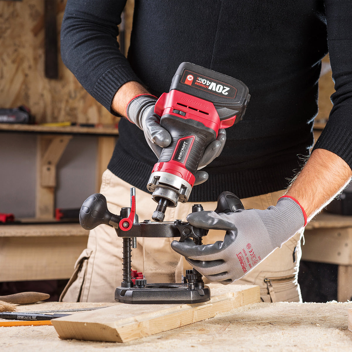 A person wearing gray work gloves uses a red and black Worcraft 20V MAX cordless router to drill a wood block in a workshop setting.