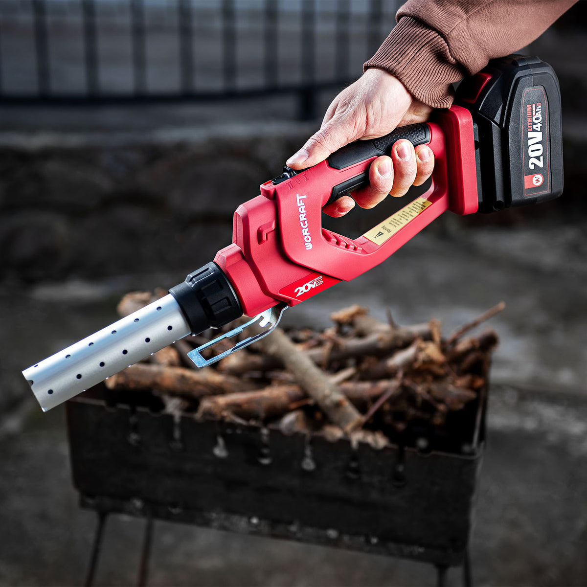 A person's hand holds a red and black Worcraft cordless firestarter over a pile of firewood logs, with a metal mouthpiece extended. The background is a dark, rustic outdoor setting.