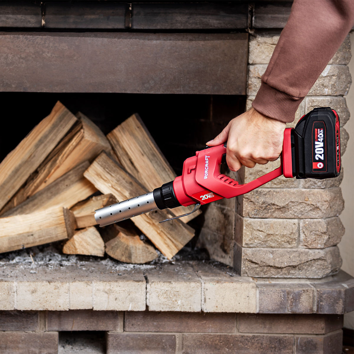 A person's hand holding a red and black cordless Worcraft fireplace starter tool in front of a stone fireplace with stacked firewood.
