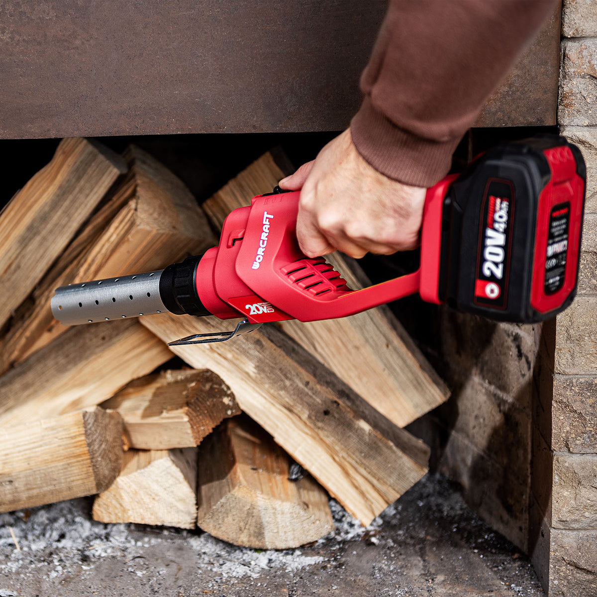 A person's hand holding a red and black Worcraft cordless firestarter over a pile of firewood logs in a fireplace opening.