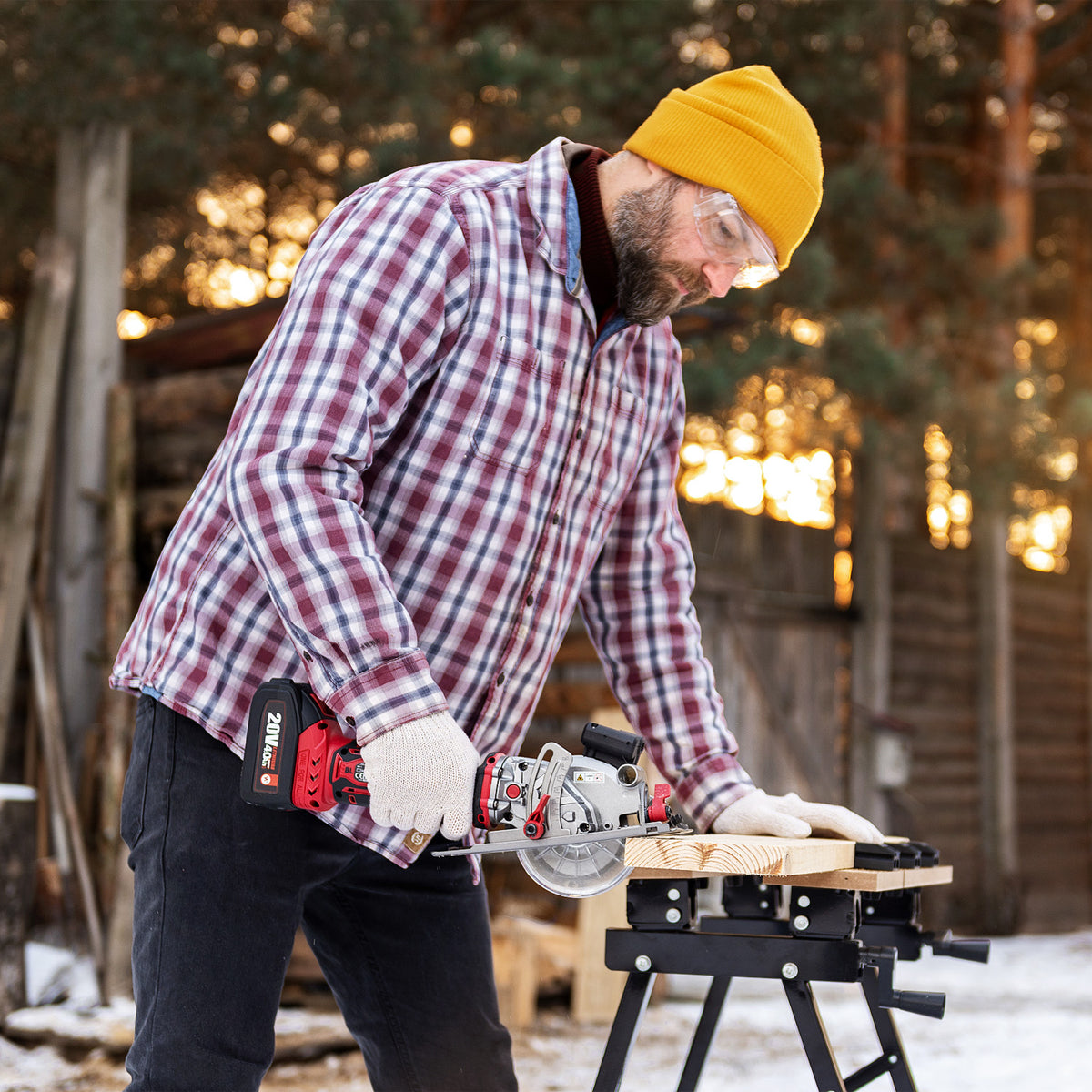 A man  uses a Worcraft mini cordless circular saw to cut a piece of wood on a black sawhorse outdoors in a snowy backyard.