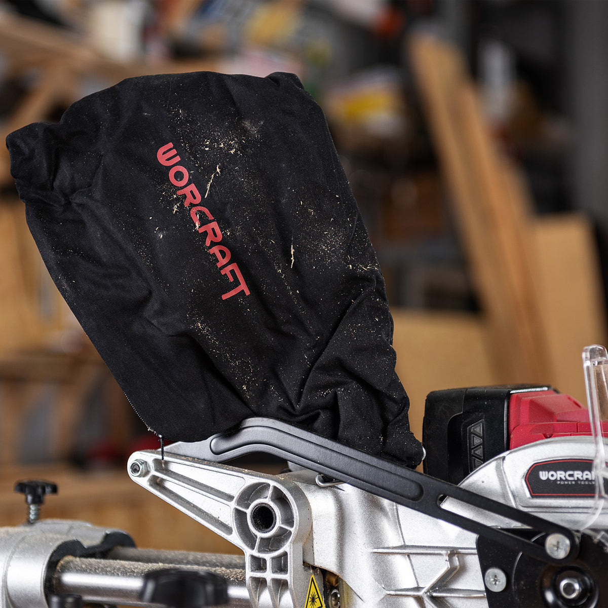 A black WorCraft dust bag with red logo lettering, shown attached to a silver and black miter saw in a workshop setting.