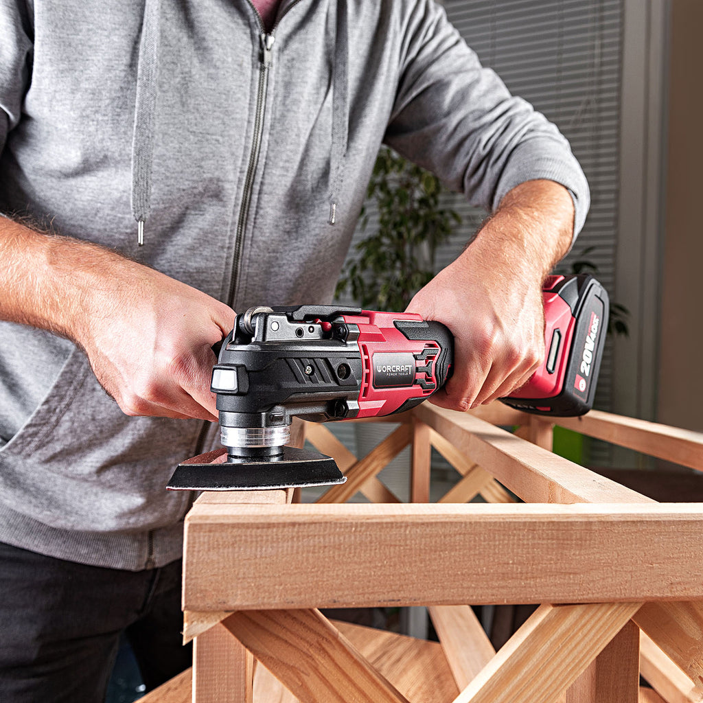 A man in a gray uses a Worcraft red and black cordless multi-tool with a sanding attachment on a wooden frame in an indoor setting.