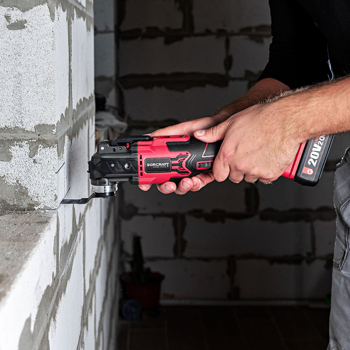 A person using a red and black Worcraft oscillating multi tool to cut a metal bracket against an unfinished concrete block wall.