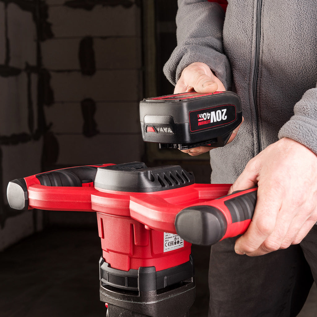 A person in a gray fleece jacket holds a Worcraft red and black cordless rotary hammer drill battery and a red-and-black cordless drill attachment and battery against a dark workshop background.
