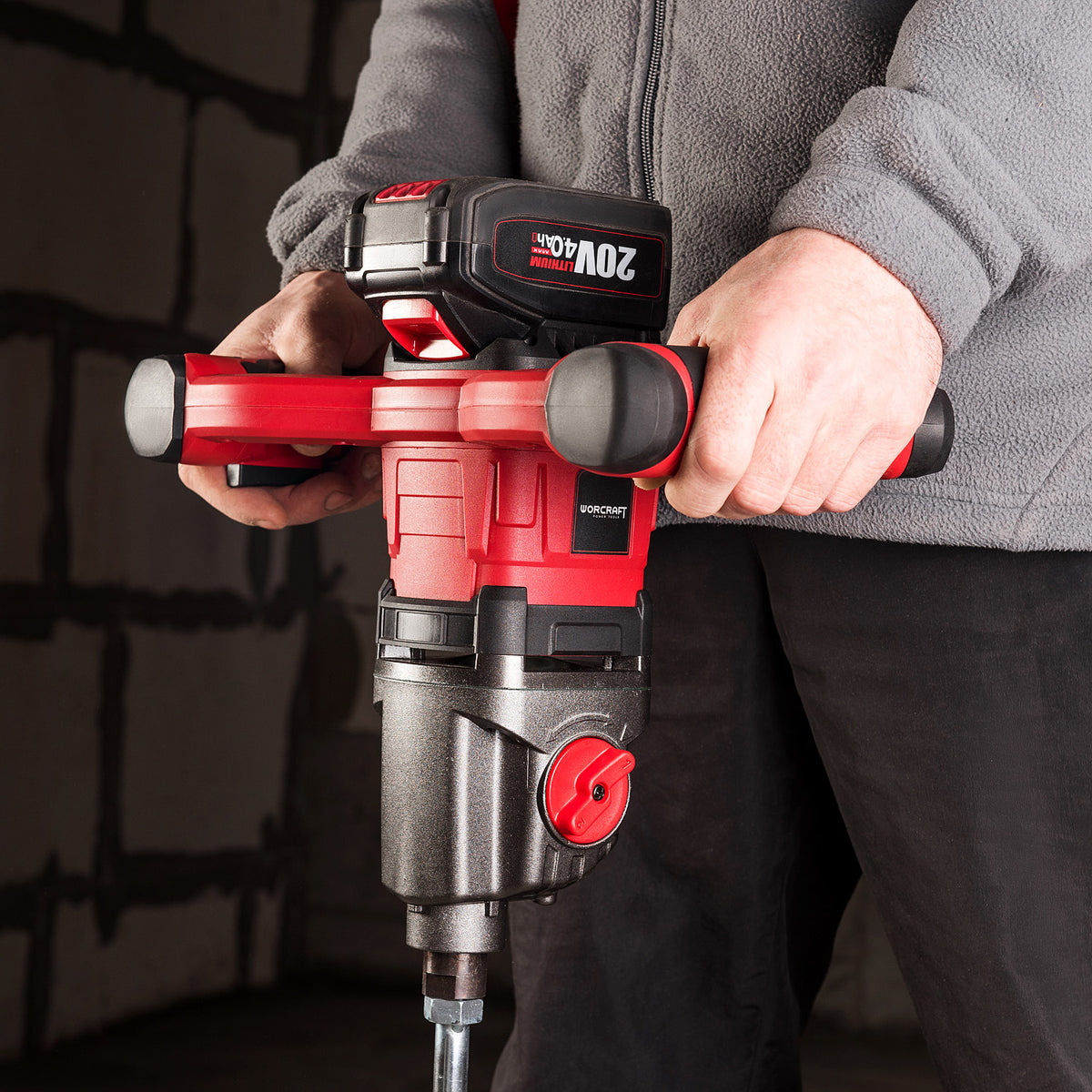 A person in a gray fleece jacket and black pants holds a red and black cordless Worcraft paddle mixer against a dark brick wall background.
