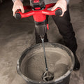 A person using a Worcraft red and black cordless drill mixer with a metal paddle attachment, mixing gray concrete or wet cement inside a large black bucket on a concrete floor.