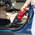 A person uses a Worcraft red and black cordless handheld car vacuum cleaner inside a blue car door and seat area. The vacuum has visible branding and the battery pack is attached.
