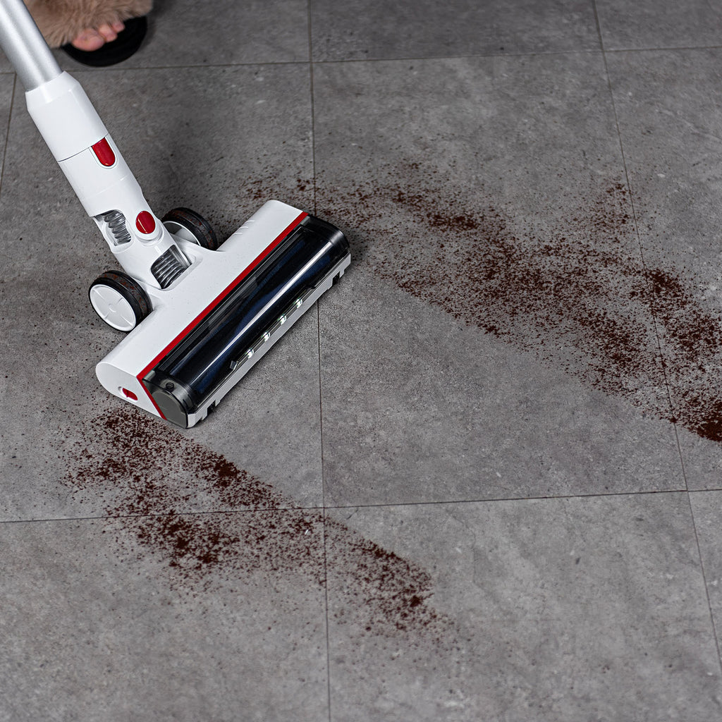 A Worcraft cordless stick vacuum cleaner with a white and red body and black roller is shown on a gray tiled floor, sucking up dark brown spilled crumbs. 