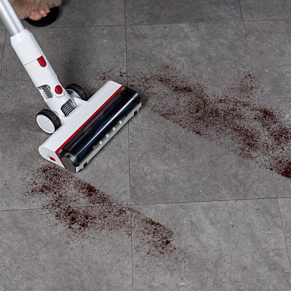 A Worcraft cordless stick vacuum cleaner with a white and red body and black roller is shown on a gray tiled floor, sucking up dark brown spilled crumbs. 
