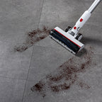 A cordless stick vacuum cleaner with a white and silver body and red accents is shown on a gray tiled floor, cleaning up dark brown debris.