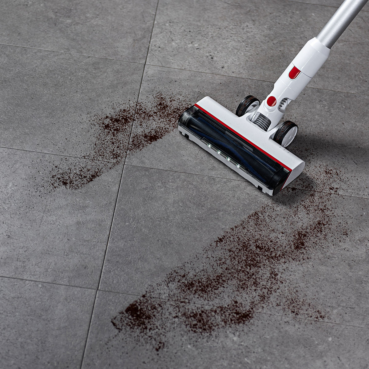A cordless stick vacuum cleaner with a white and silver body and red accents is shown on a gray tiled floor, cleaning up dark brown debris.