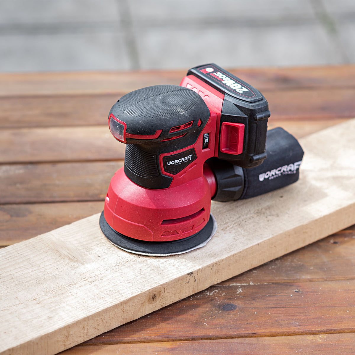 A red and black WorCraft cordless orbital sander resting on a light-colored wooden board over a wooden workbench.