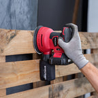 A red and black Worcraft cordless orbital sander being used by a hand wearing a gray work glove to sand a wooden pallet.