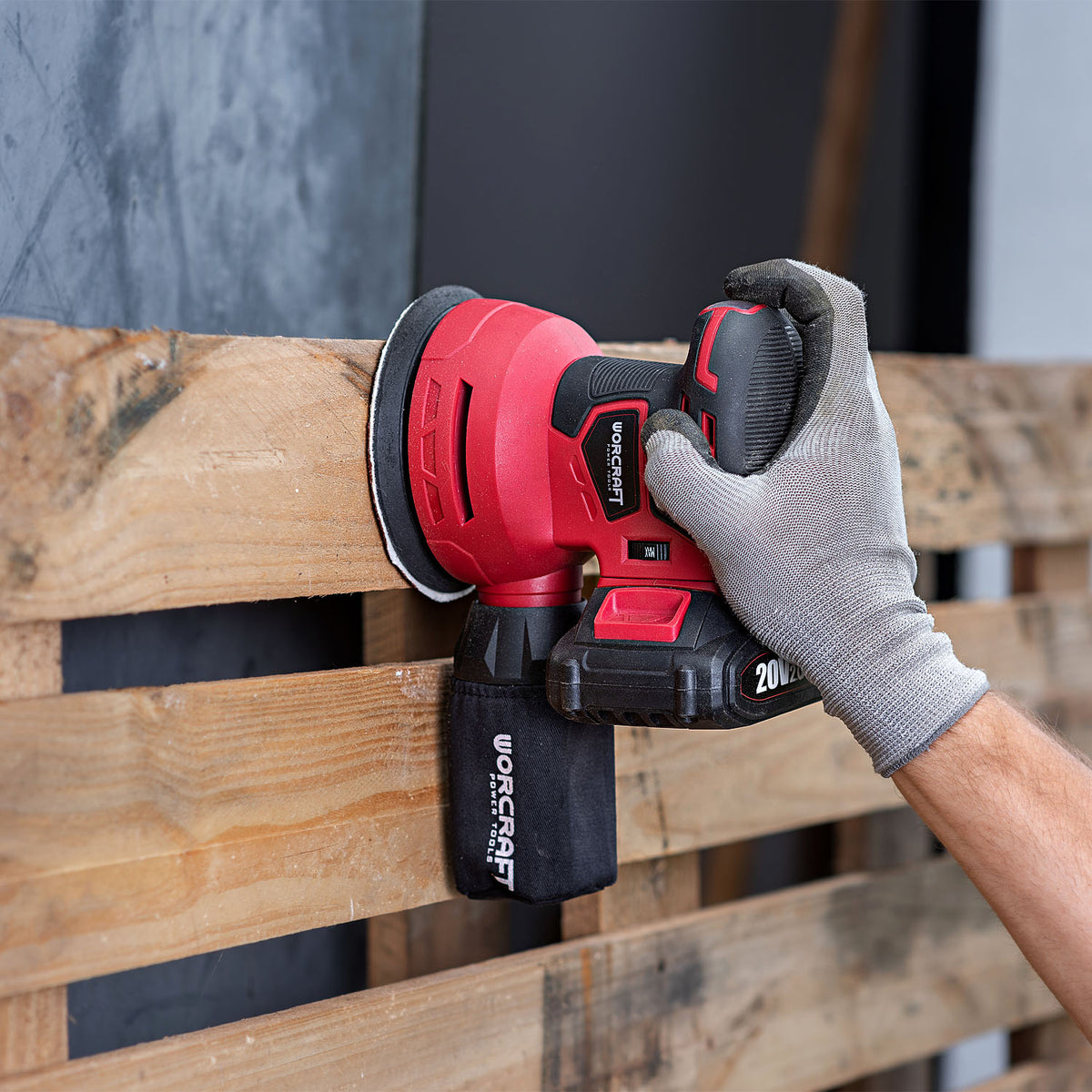 A red and black Worcraft cordless orbital sander being used by a hand wearing a gray work glove to sand a wooden pallet.