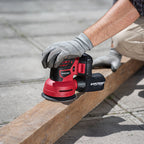 A person wearing gray work gloves uses a red and black cordless orbital sander on a wooden beam outdoors on concrete pavement. The brand name 'WORCRAFT' is clearly visible on the tool.