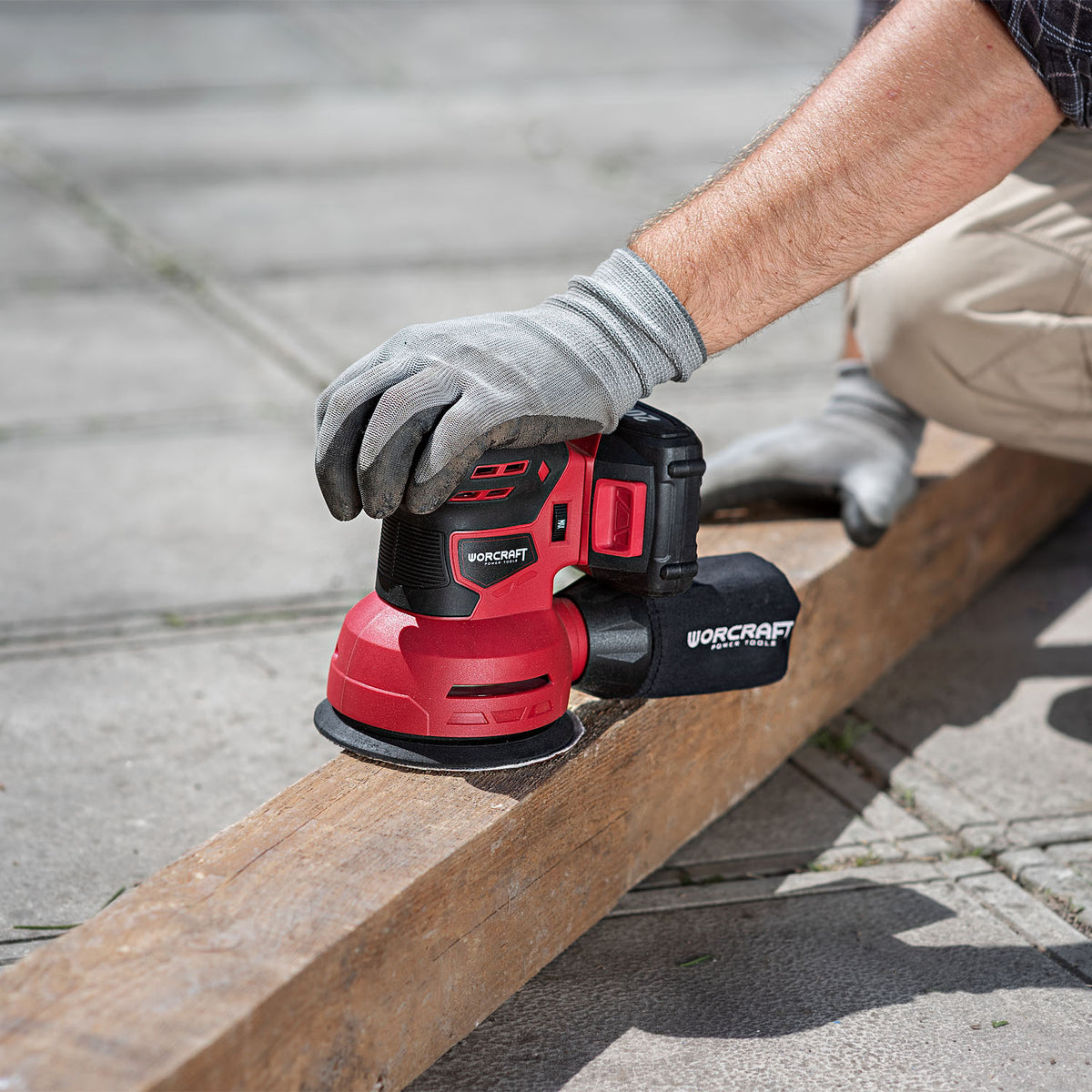 A person wearing gray work gloves uses a red and black cordless orbital sander on a wooden beam outdoors on concrete pavement. The brand name 'WORCRAFT' is clearly visible on the tool.