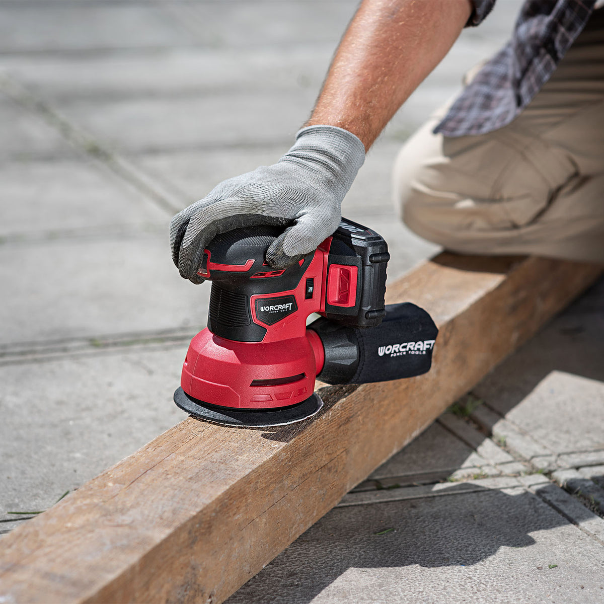 A person wearing a gray work glove uses a red and black Worcraft cordless orbital sander to smooth a wooden beam outdoors on concrete pavement.