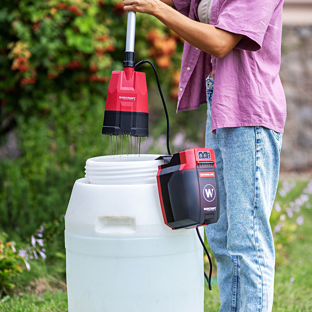 A person outdoors uses a red and black Worcraft cordless  attachment with a long metal wand to mix contents in a large white plasticrain barrel. 