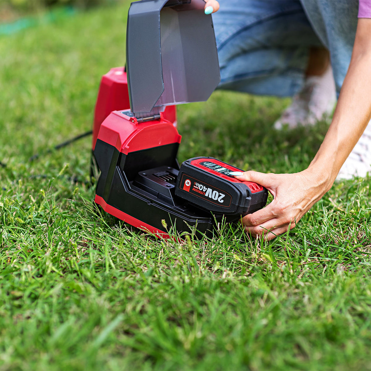 A person kneeling on green grass charges a red and black cordless battery with a red and black charging dock. The battery has a 20V 18Ah label.