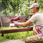 A man in a beige work uniform and cap uses a red Worcraft cordless paint sprayer to apply paint to a wooden swing in an outdoor garden setting.