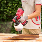A person wearing a white and red shirt, gray gloves, and khaki pants uses a red and black Worcraft cordless paint sprayer to apply a brown wood stain to a wooden table outdoors.