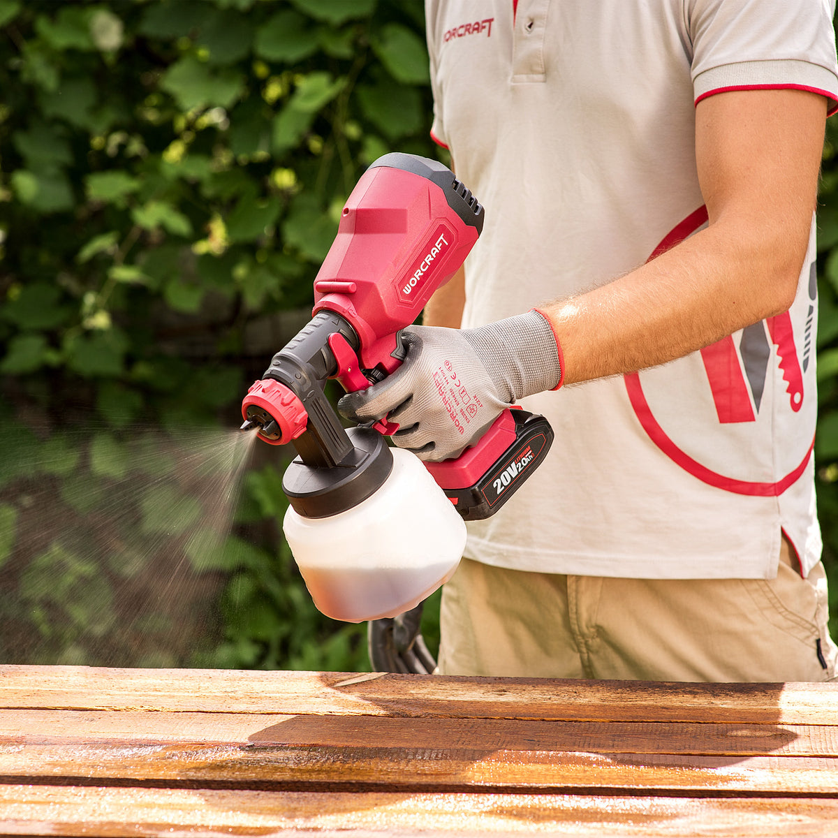 A person wearing a white and red shirt, gray gloves, and khaki pants uses a red and black Worcraft cordless paint sprayer to apply a brown wood stain to a wooden table outdoors.