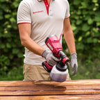 A person wearing a white and red Worcraft polo shirt and gray gloves operates a red and black Worcraft cordless paint sprayer over a wooden surface, with a blurred green garden background.