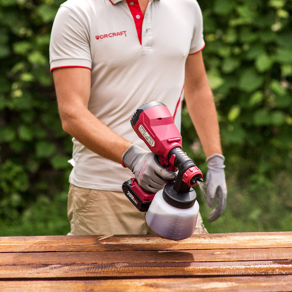 A person wearing a white and red Worcraft polo shirt and gray gloves operates a red and black Worcraft cordless paint sprayer over a wooden surface, with a blurred green garden background.