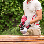A person wearing a beige and red utility polo shirt and gray work gloves holds a red and black Worcraft cordless paint sprayer over a wooden surface, with a blurred green garden background.