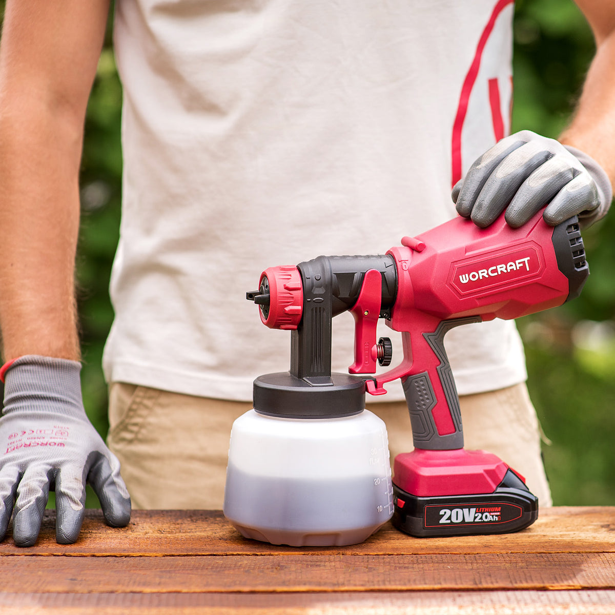 A person wearing work gloves holds a red and black Worcraft cordless paint sprayer with a paint container on a wooden table outdoors.