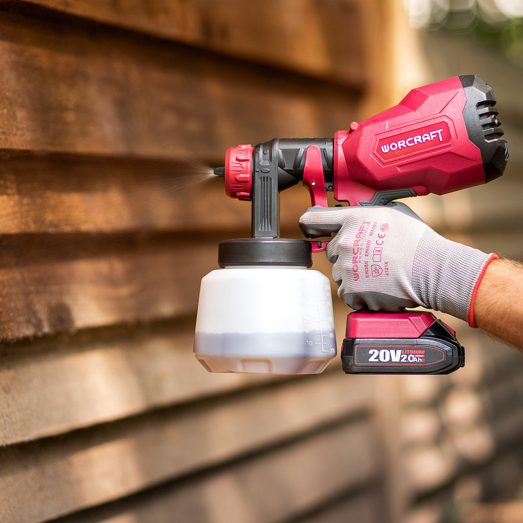 A person wearing a gray work glove holds a red and black Worcraft cordless paint sprayer against a blurred wooden siding background.