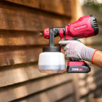 A person wearing a gray work glove holds a red and black Worcraft cordless paint sprayer against a blurred wooden siding background.