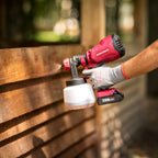 A person wearing a gray work glove uses a red and black Worcraft cordless paint sprayer to apply wood stain to a wooden fence. The Gorcraft brand logo is clearly visible on the tool, and a 20V 14.