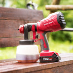 A red and black WORCRAFT cordless paint sprayer gun with a clear paint container, resting on a wooden deck with a blurred garden background.