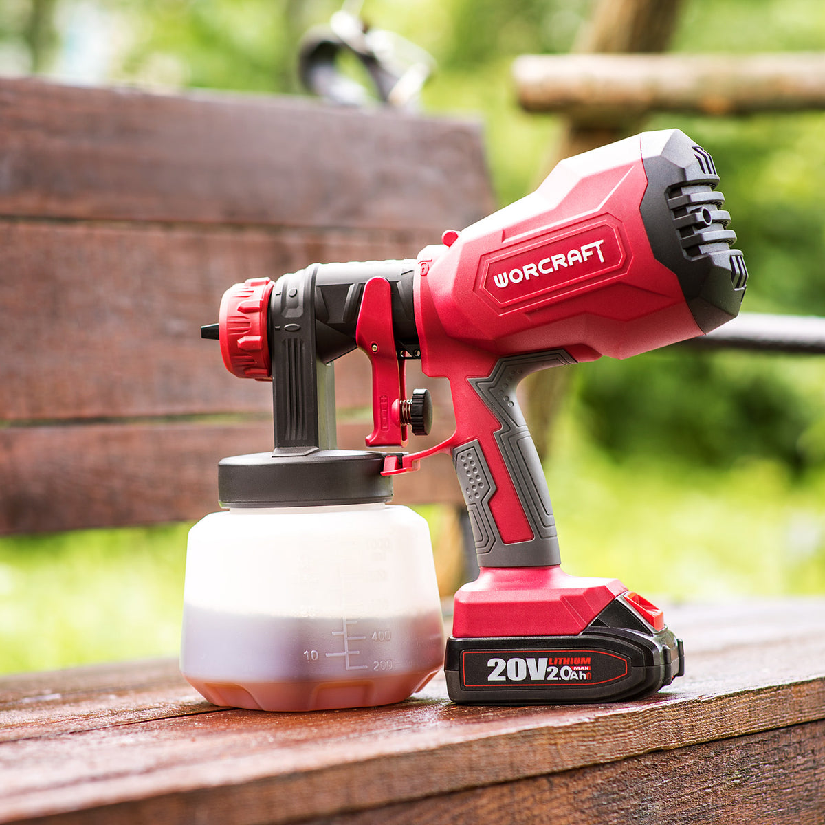 A red and black WORCRAFT cordless paint sprayer gun with a clear paint container, resting on a wooden deck with a blurred garden background.