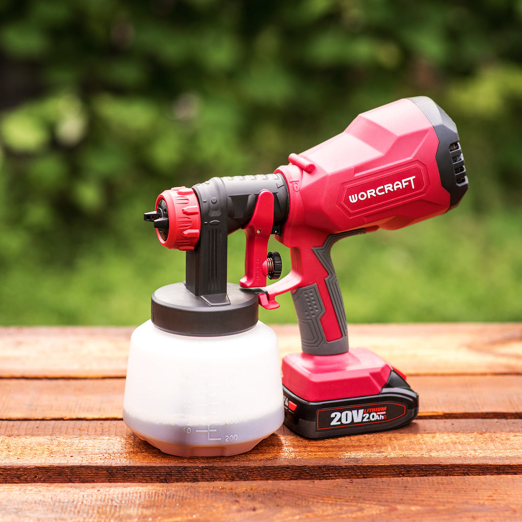 A red and black WorCraft cordless paint sprayer with a white plastic paint container, resting on a wooden deck with a blurred green garden background.
