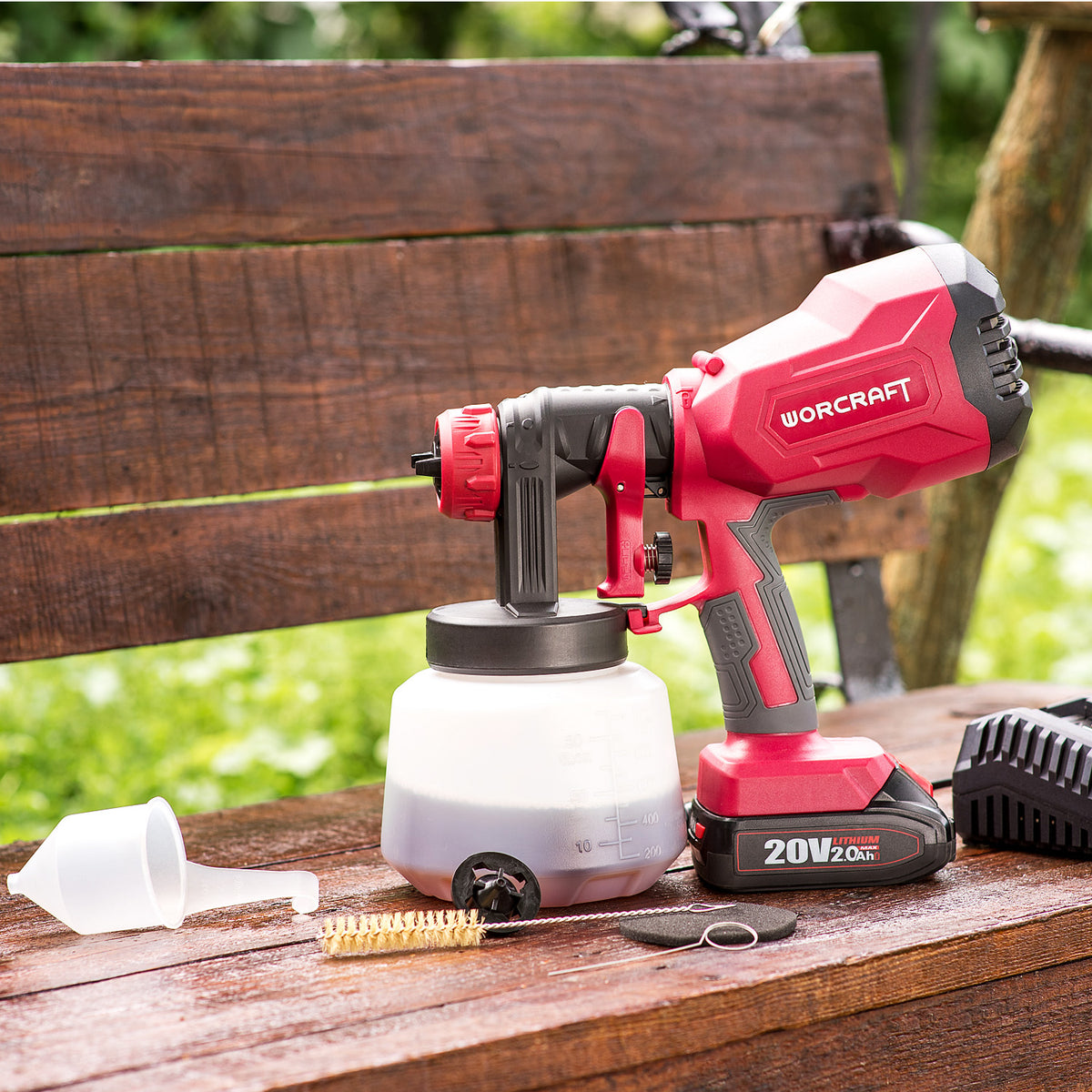 A red and black Worcraft 20V 18V cordless paintball gun with a white paint container, set on a wooden bench outdoors with a paint brush, funnel, and battery charger nearby.
