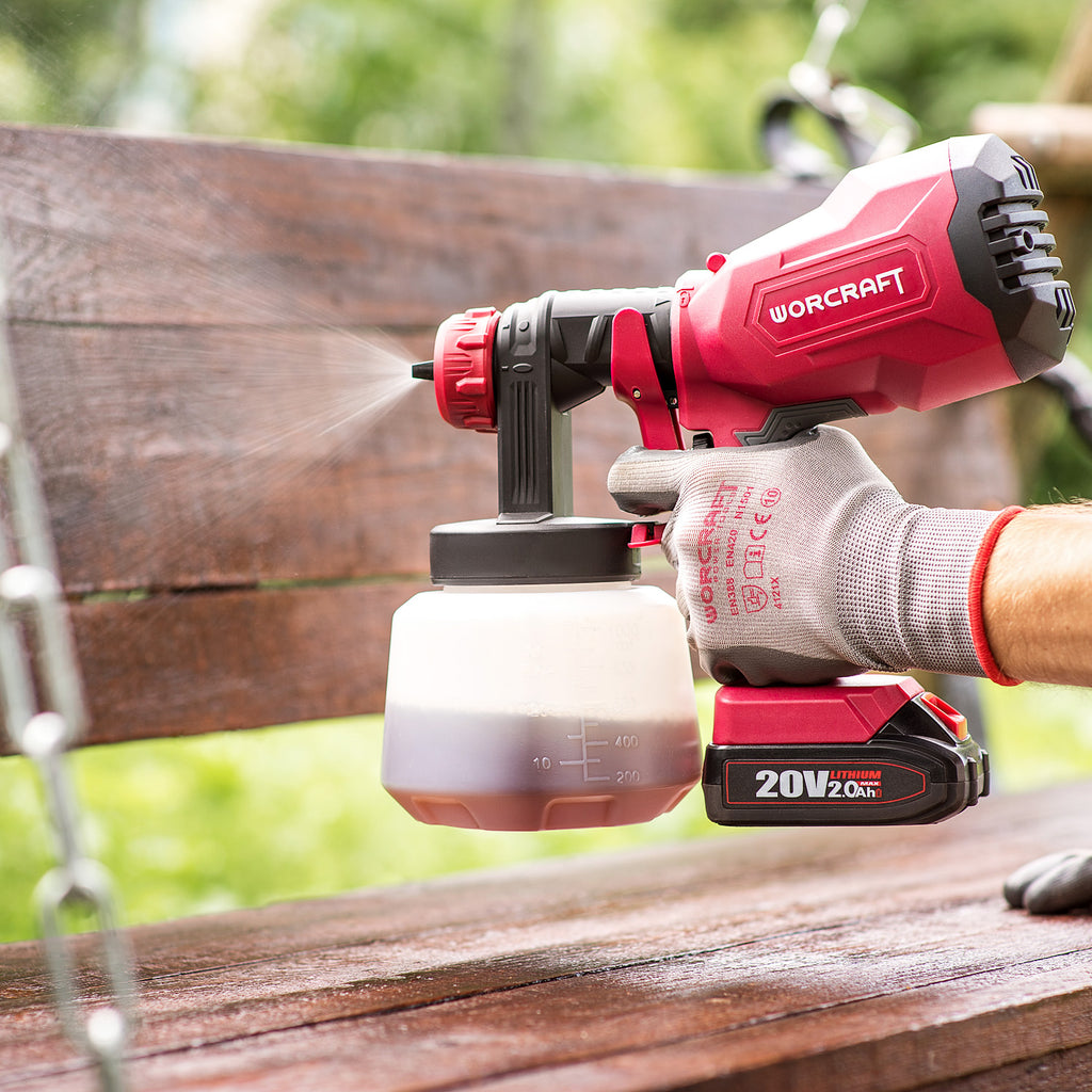 A red and black Worcraft cordless paint sprayer being used by a gloved hand to spray a wooden bench outdoors. The spray nozzle is directed at the wood, with a visible mist and a white fuel container below.