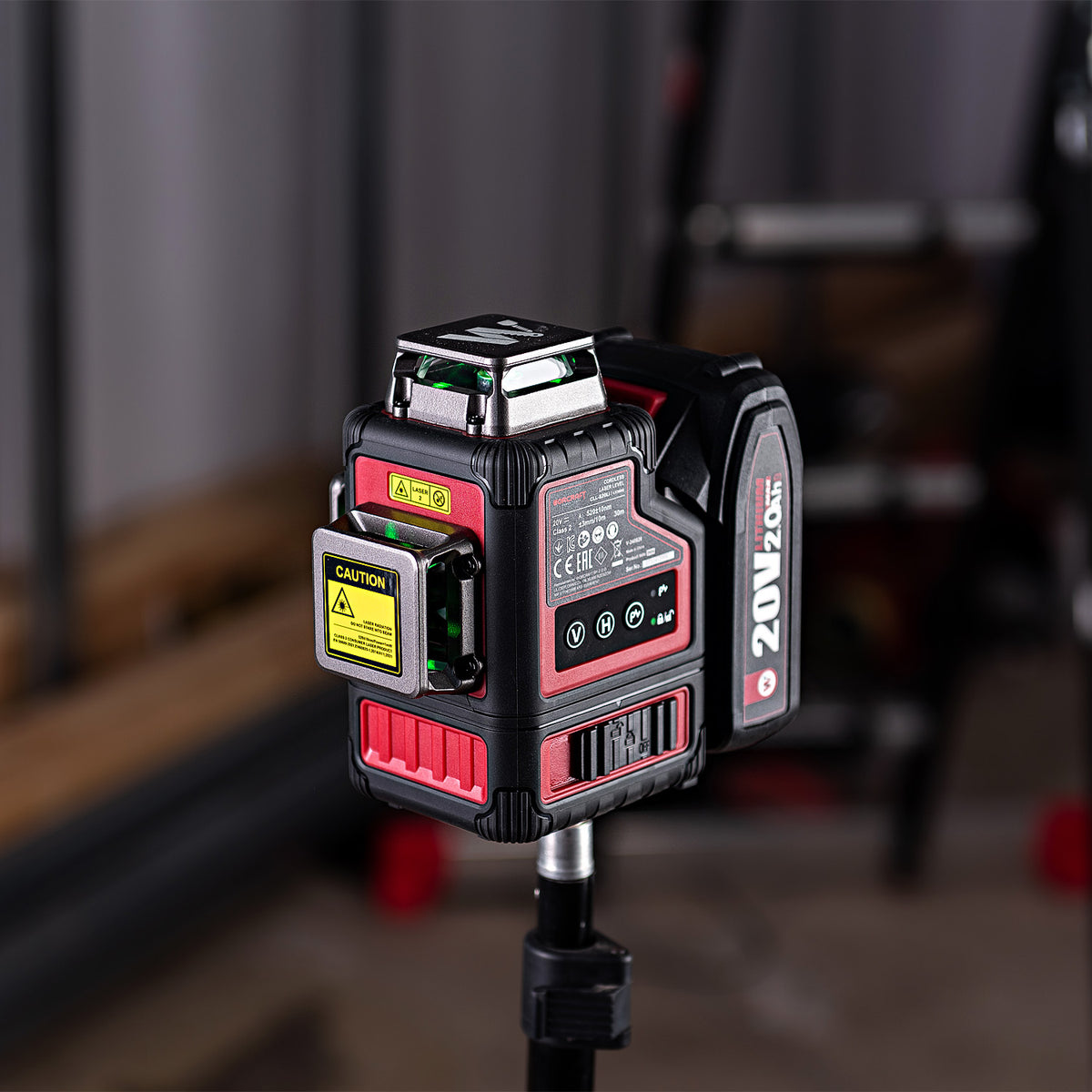 A red and black 20V 20W laser level mounted on a tripod, shown in a workshop setting with a blurred background.