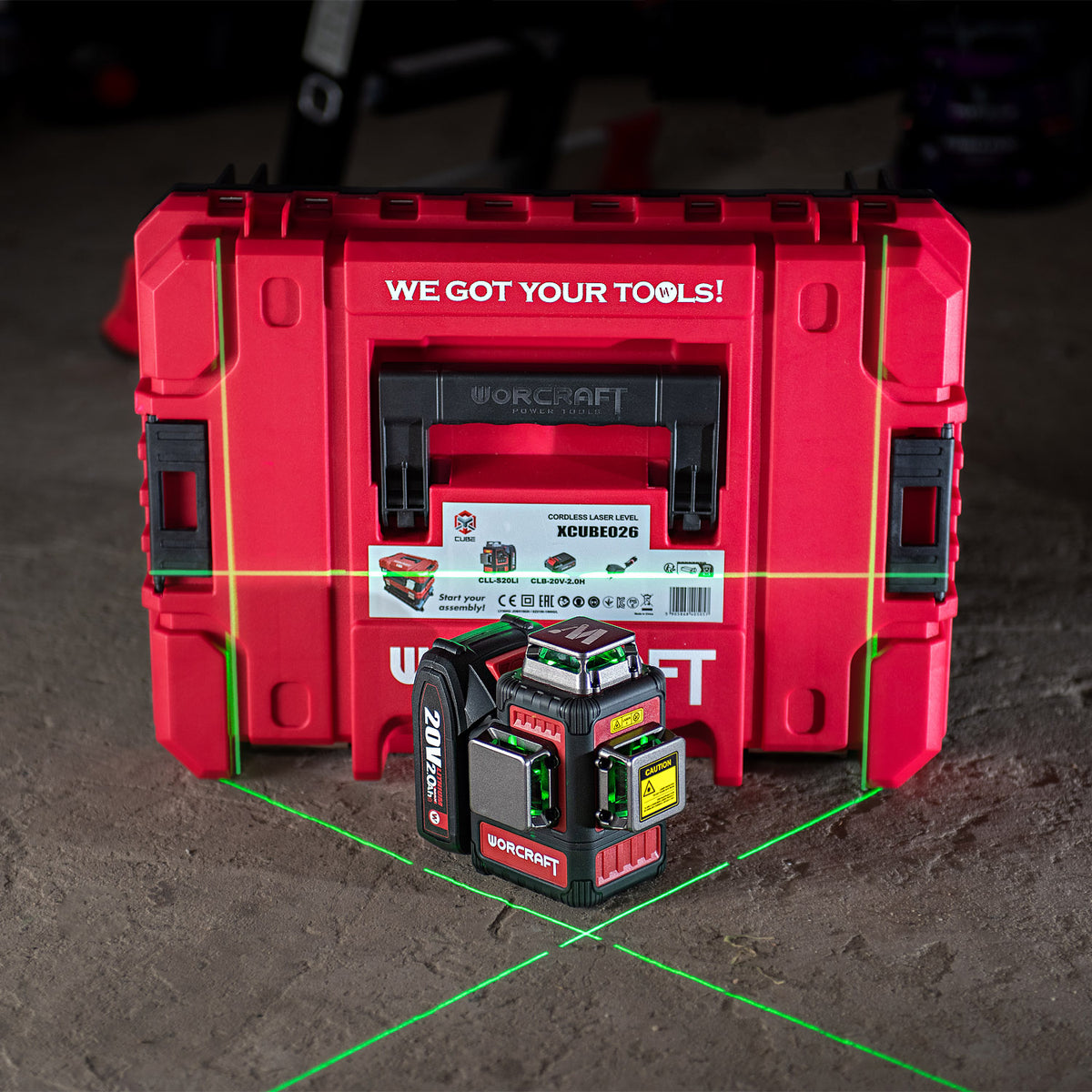A red Worcraft Workstation carrying case with a black and red Worcraft XCUBE026 K20 Pro laser level placed in front of it on a concrete floor. Green and red laser lines are visible on the floor, and the background is a dark workshop environment.