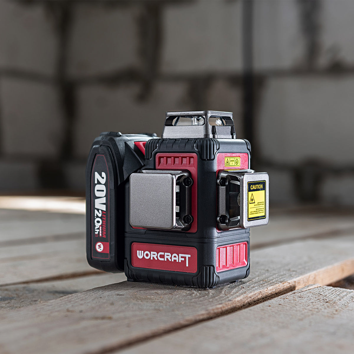 A Worcraft 20V 2.0Ah cordless laser level device in black and red, shown with its battery attached on a wooden surface with a blurred concrete background.